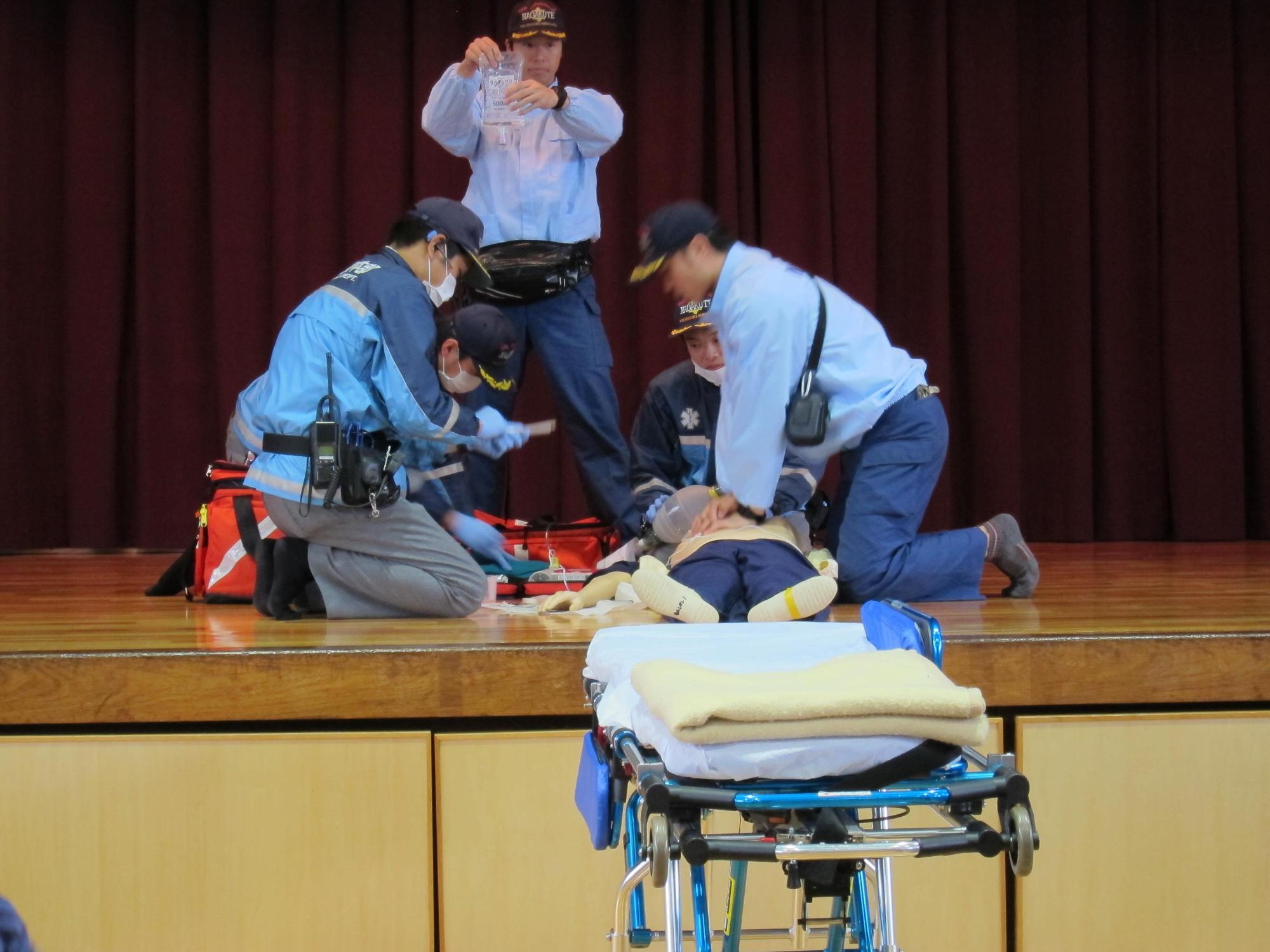 体育館のステージで人形を使って消防署員による救急活動訓練が行われている前に担架が置いてある写真