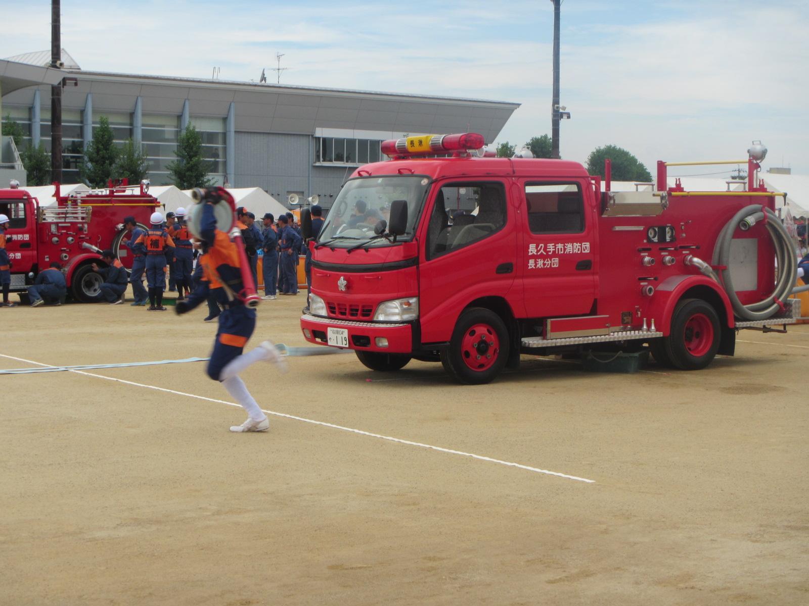 運動場の消防車両の前を団員が放水ホースを担いで走っている写真