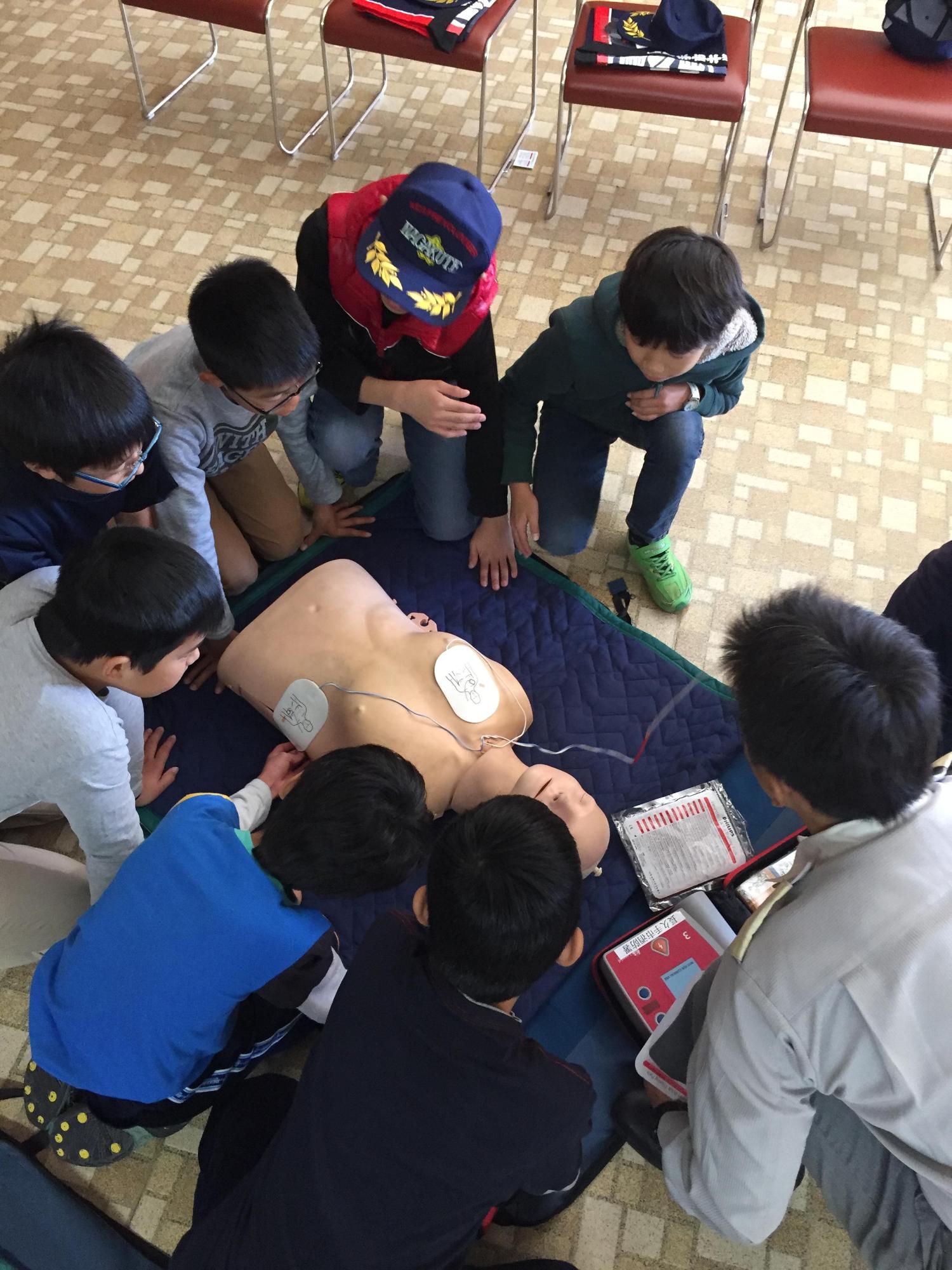 救命講習にてAEDが貼られた人形を見ながら救急隊員の説明を聞いているキッズ団員たちの写真