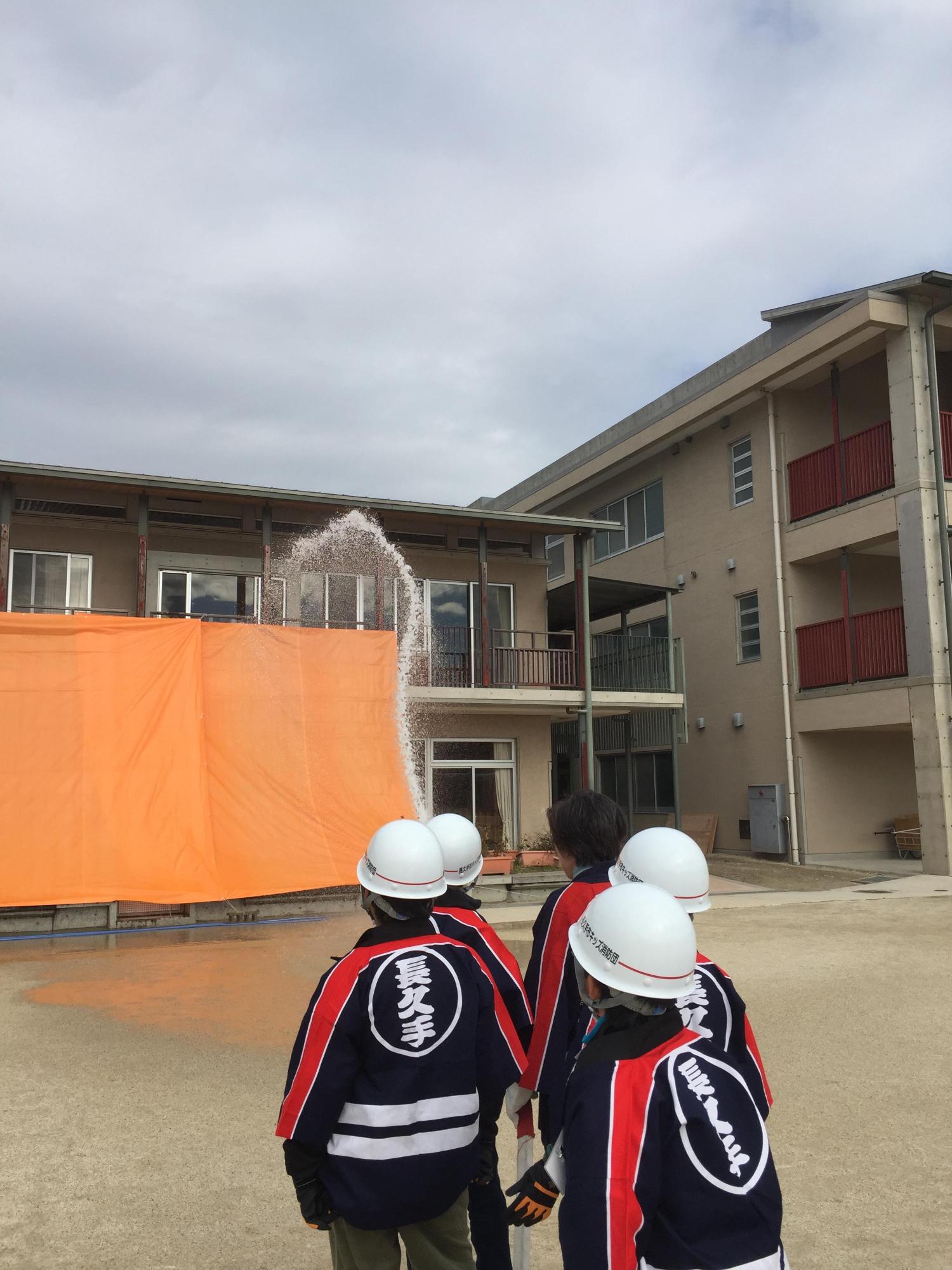 建物に張ってあるオレンジのシートをめがけてヘルメットに法被姿のキッズ消防団員たちが放水作業している写真