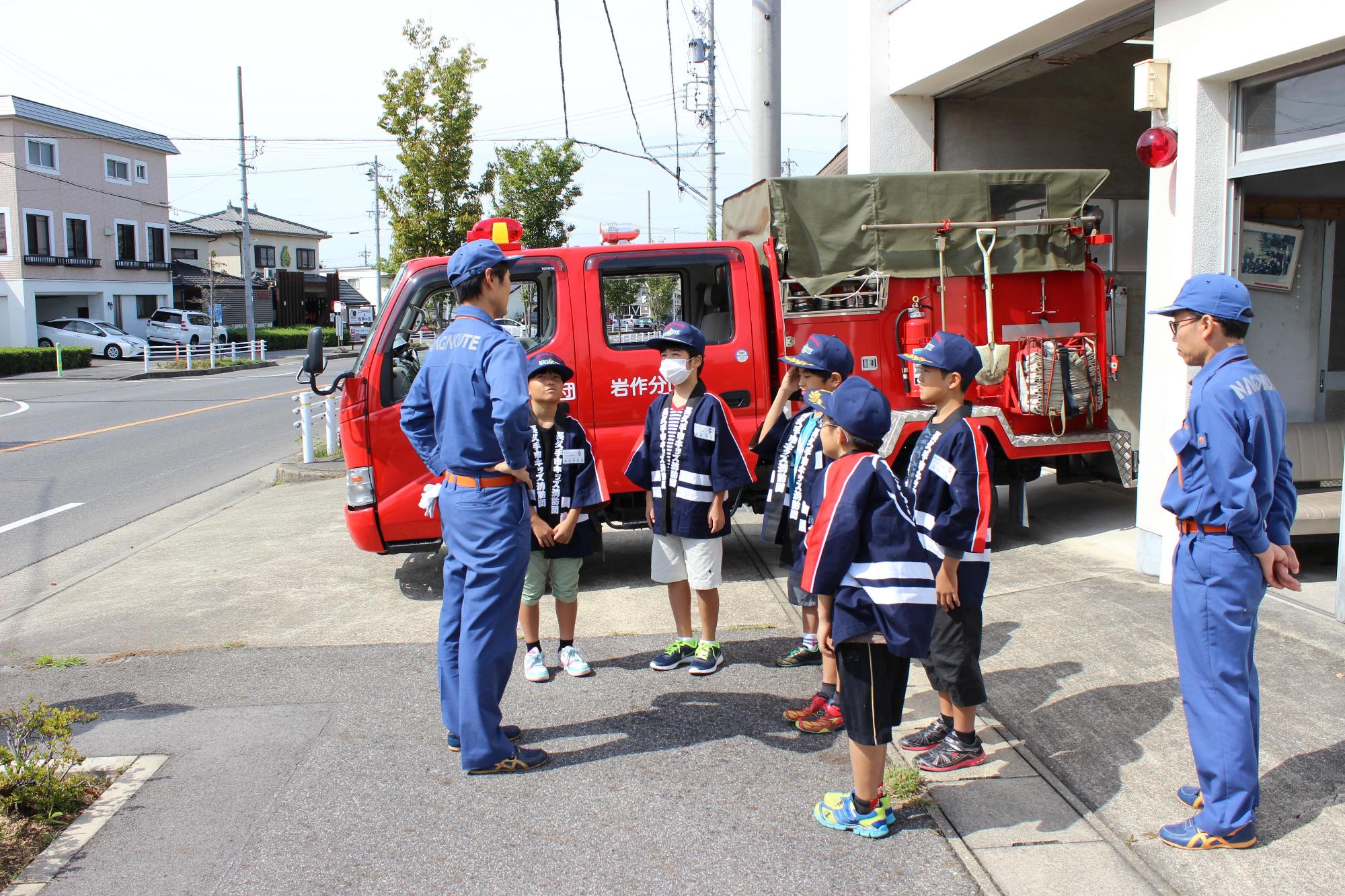 消防車両の前で団器具庫の説明を聞くキッズ消防団員たちの写真