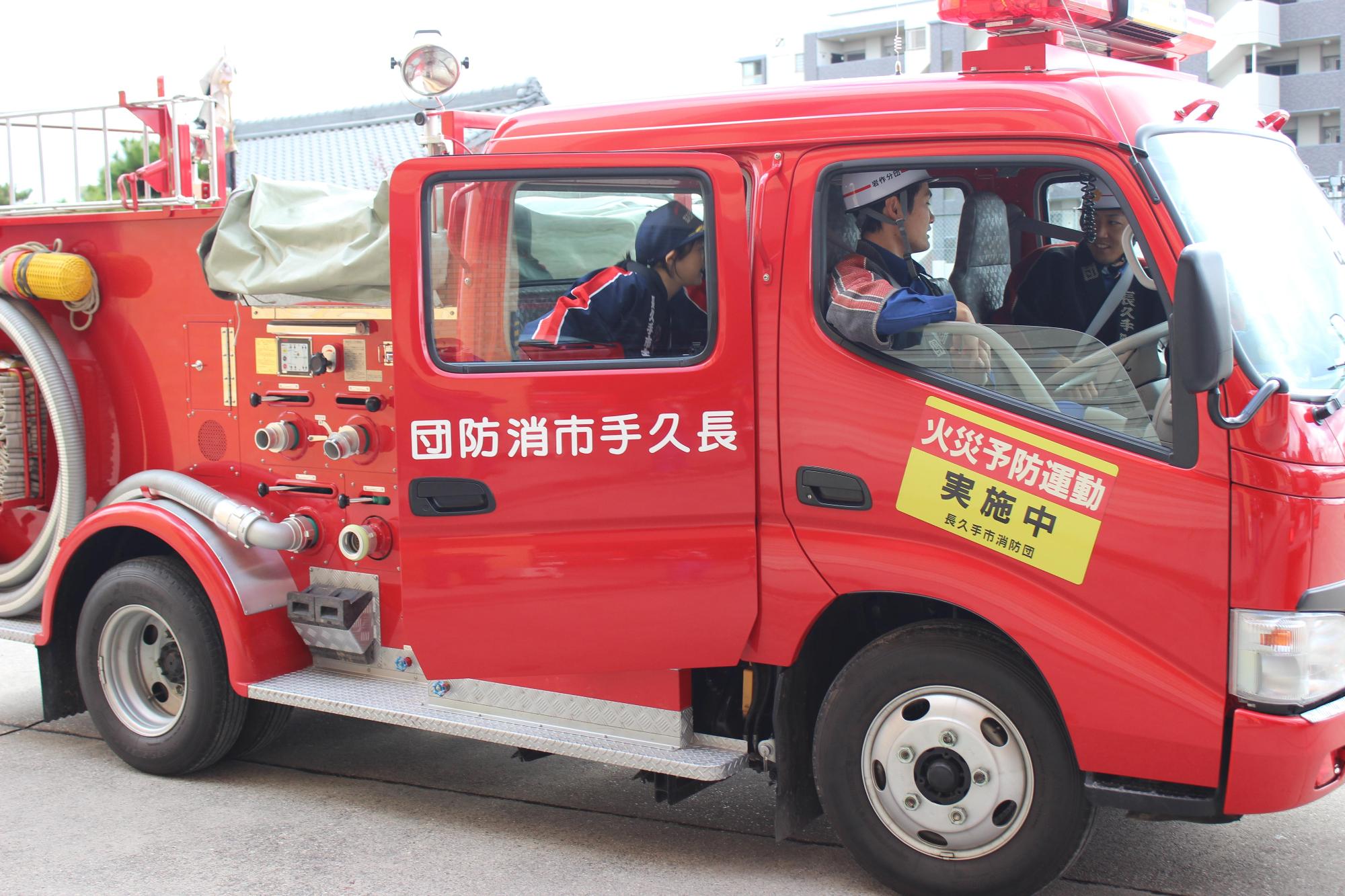 消防団員2名が消防車両の運転席と助手席に乗っており、後部座席に乗り込むキッズ団員の写真