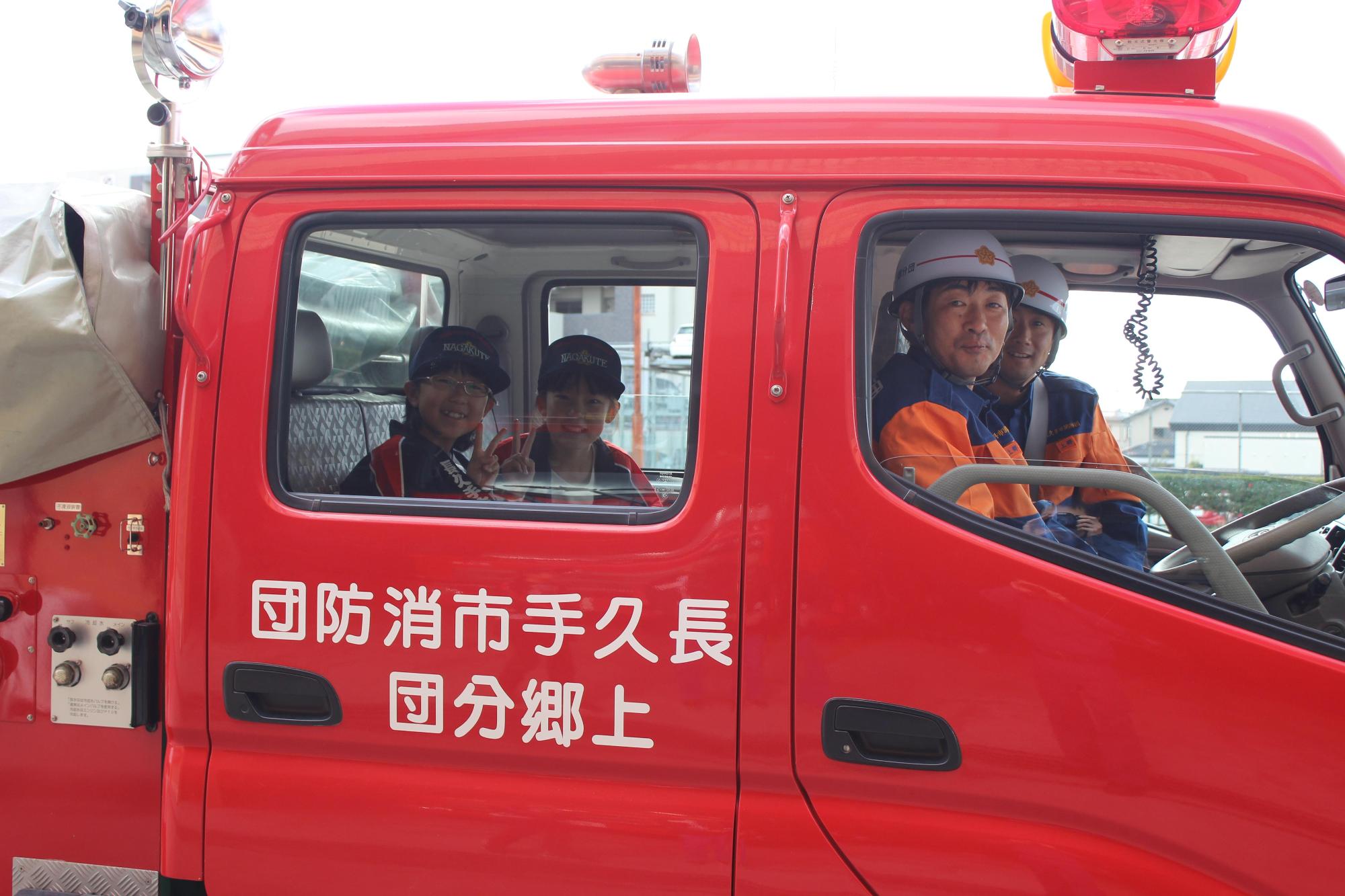 消防団員2名が消防車両の運転席と助手席に乗っており、後部座席に乗ったキッズ団員2名がピースサインをしている写真
