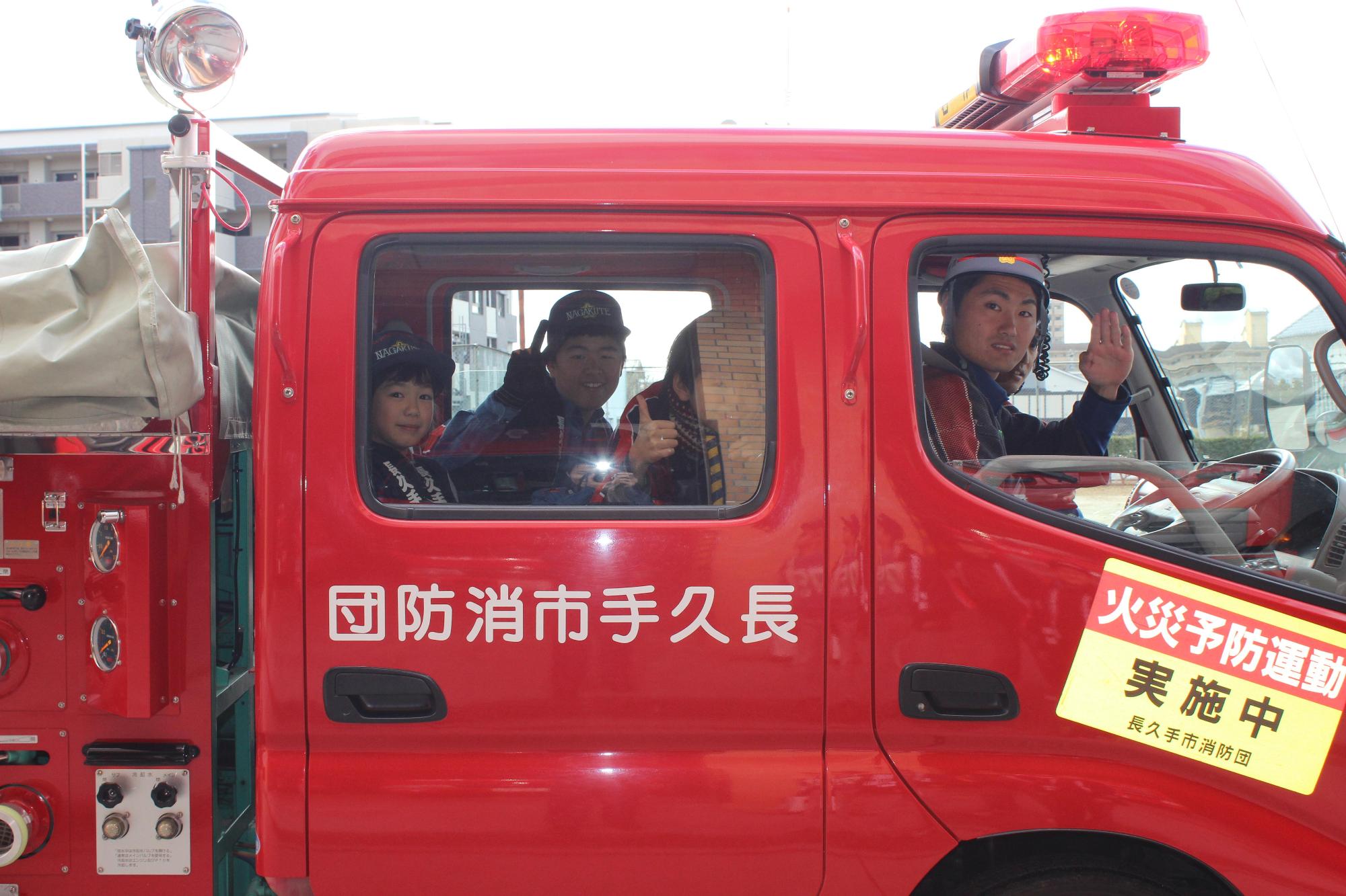 防火広報のため、消防車両を運転している消防団員が敬礼を、後部座席に乗ったキッズ団員3名がピースなどのポーズをとっている写真