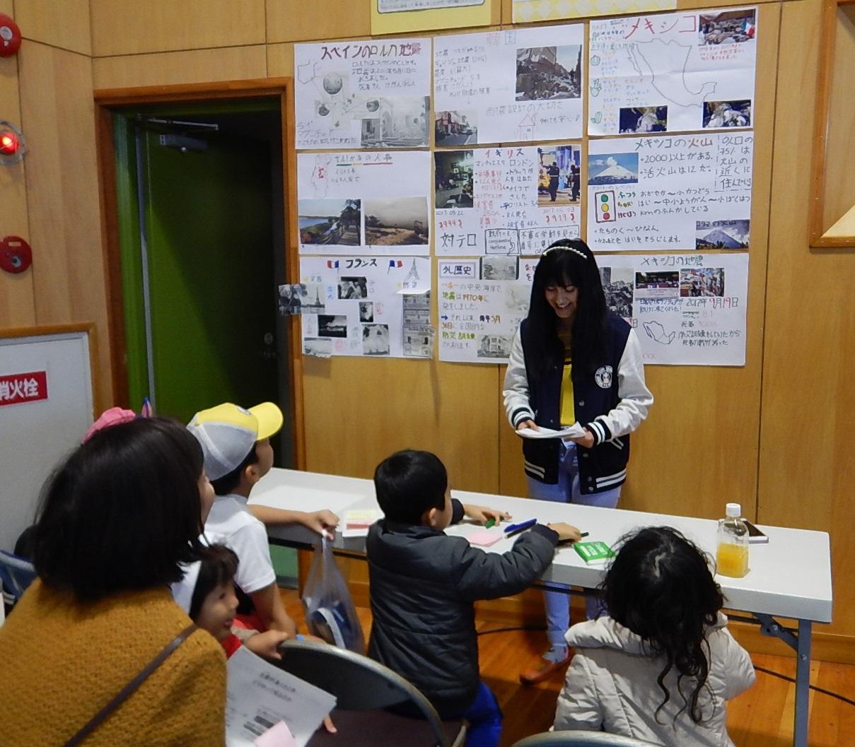 壁に各国の防災情報の紙が貼られ、参加している子供たちや保護者が壁の前に立っている女性の話を聞いている写真