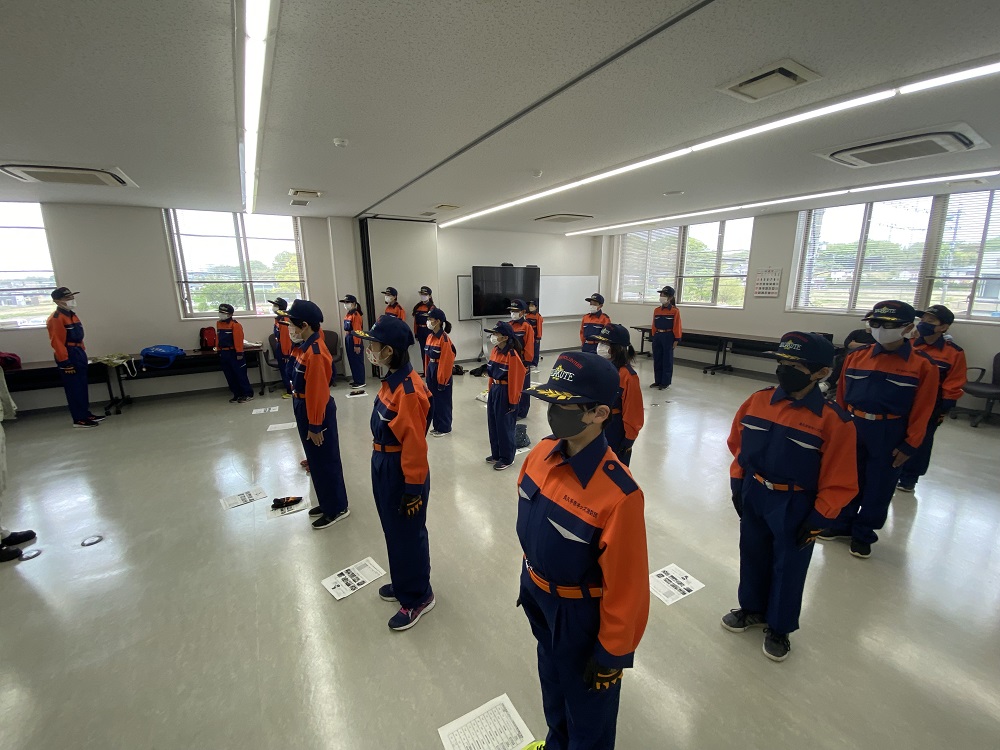 講堂の前の方でオレンジ色のベストを着た消防団員と、後ろに立って並んでいる法被姿やユニホーム姿のキッズ消防団員たちが敬礼している写真