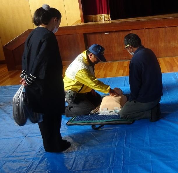 1人の女性が見守る中、青い帽子を被り黄色い上着を着た男性がもう一人の男性に人命救助人形を使い救急救命訓練をしている様子の写真