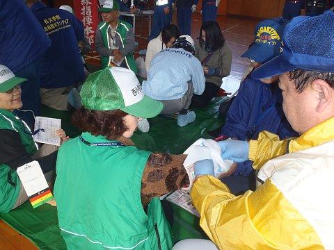 緑色のベストにキャップを被った女性の右腕に、男性ボランティアが包帯を巻いている応急手当訓練の様子の写真
