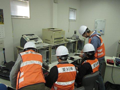 愛知県を書かれているオレンジ色のベストを着用し白色のヘルメットを被った4名のスタッフが、災害情報伝達訓練を行っている写真
