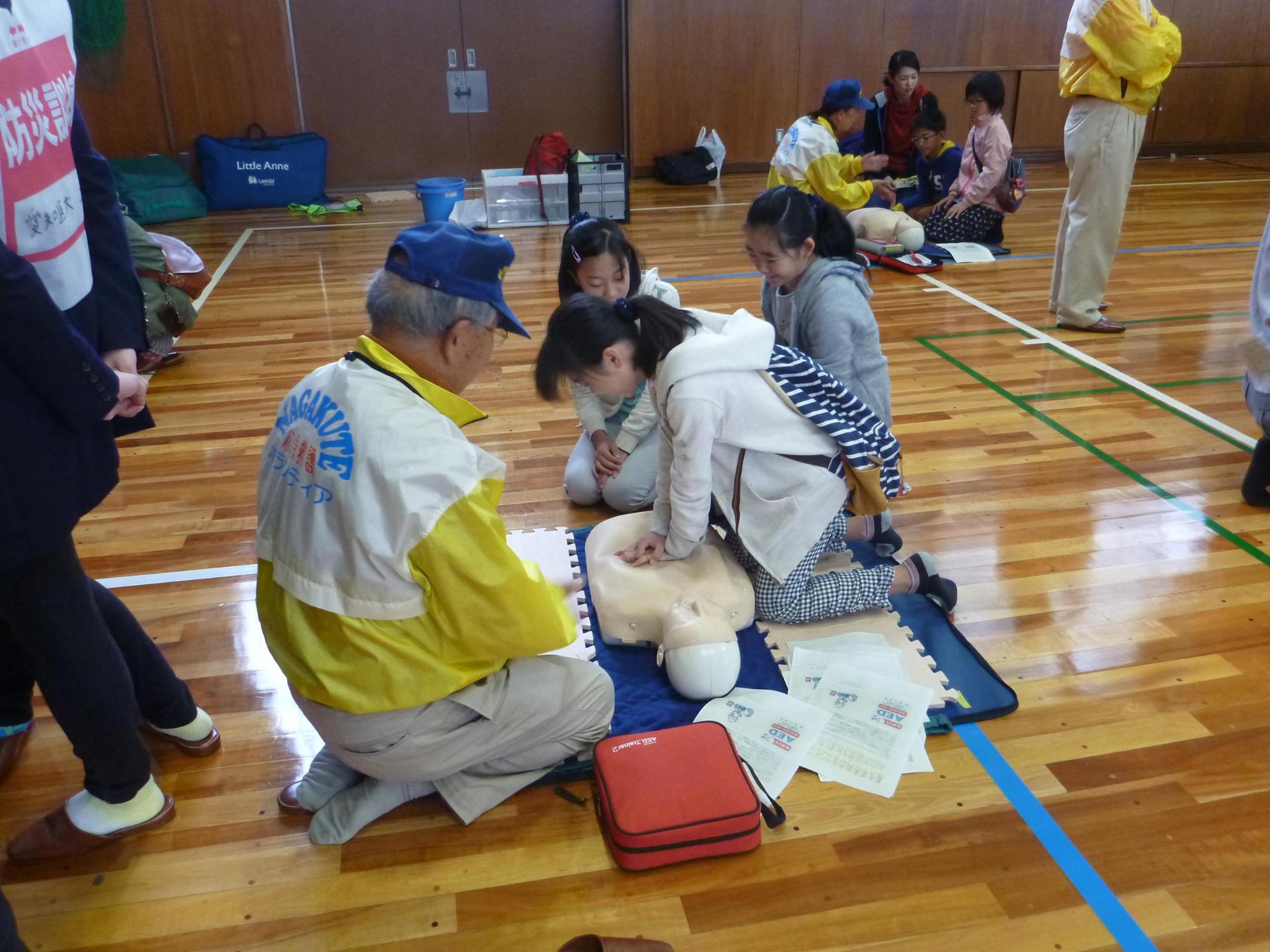 体育館にて参加者がボランティアスタッフの指導の下、AED操作訓練で人形を使って心臓マッサージをしている写真