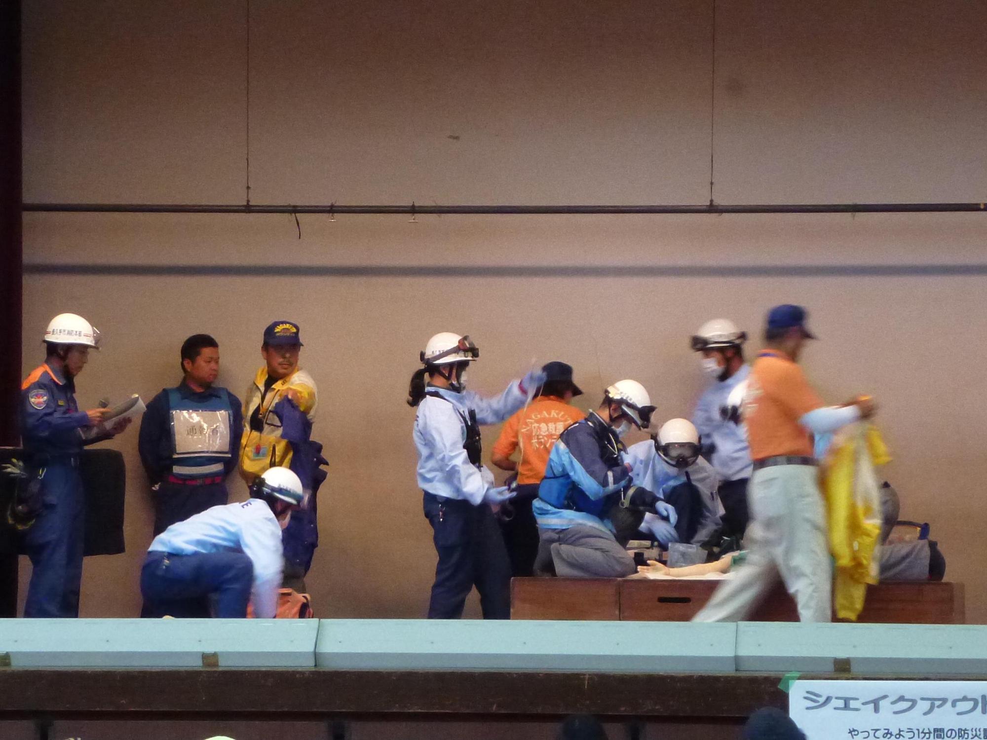 体育館のステージでヘルメットにゴーグル、マスクをした消防署員が救急活動訓練をしている写真