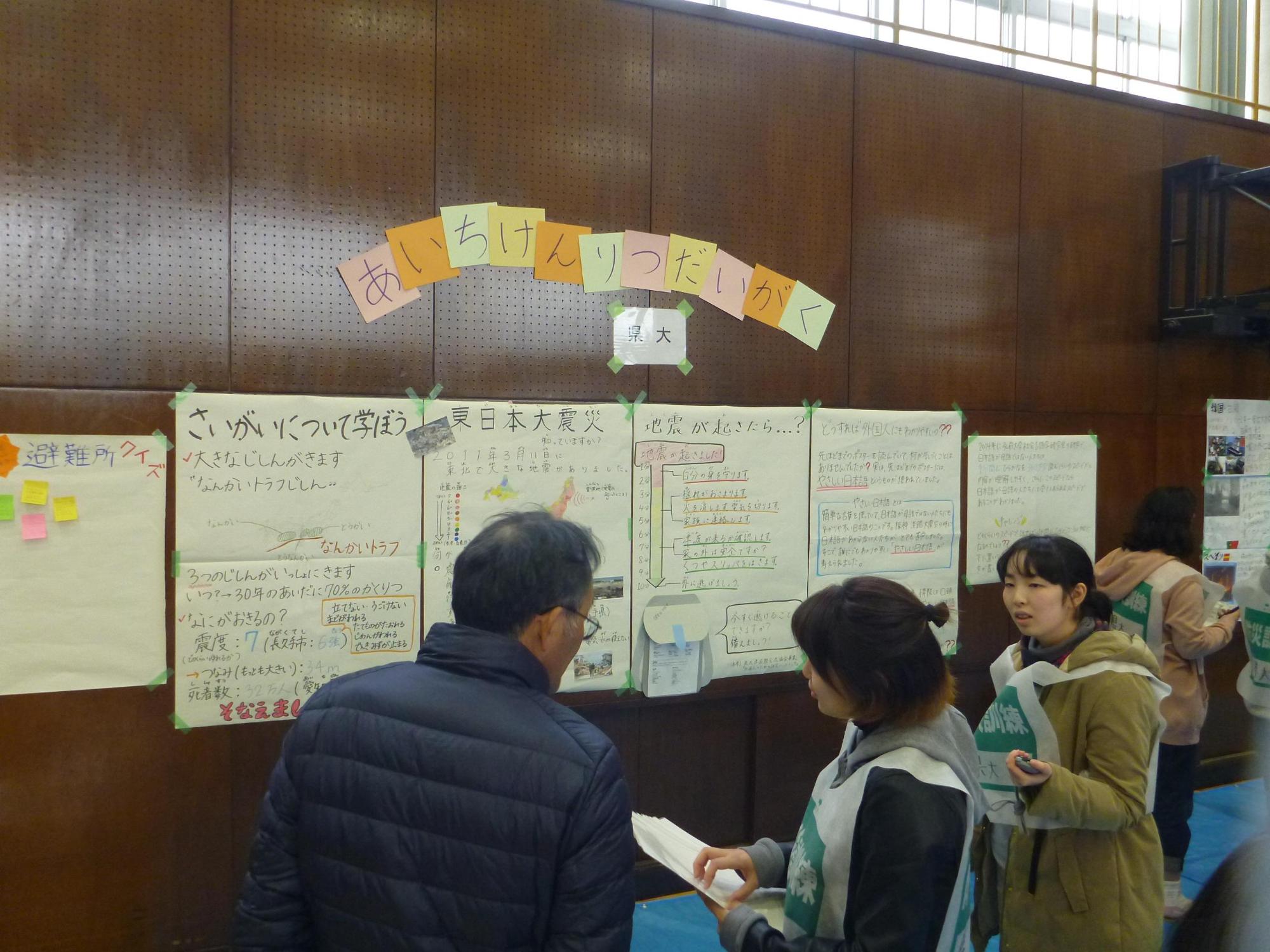 体育館の壁に避難所に関する資料がの掲示されており、参加者たちが話をしながら見学している写真