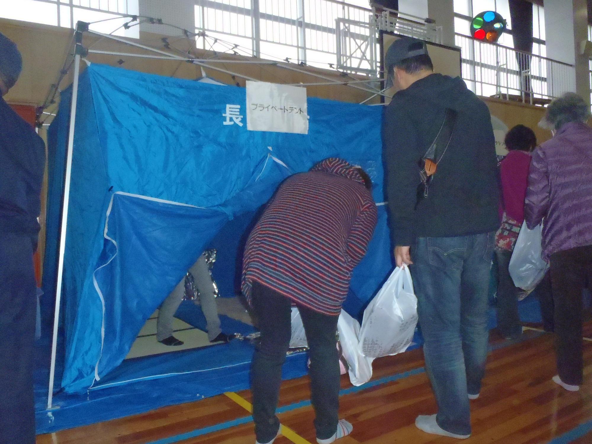 体育館に設置された青いプライベートテント(避難所)に入ろうとする参加者たちの写真