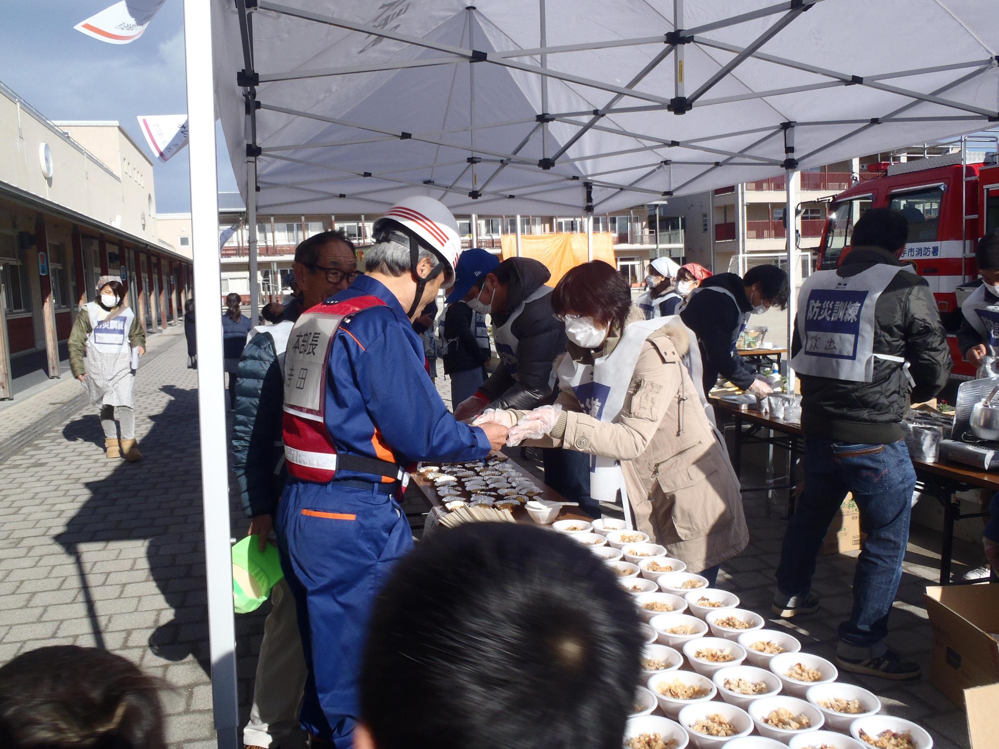炊き出しコーナーのテーブルにご飯やお菓子が並べられ、ヘルメットを被った役員が食料を受け取っている写真