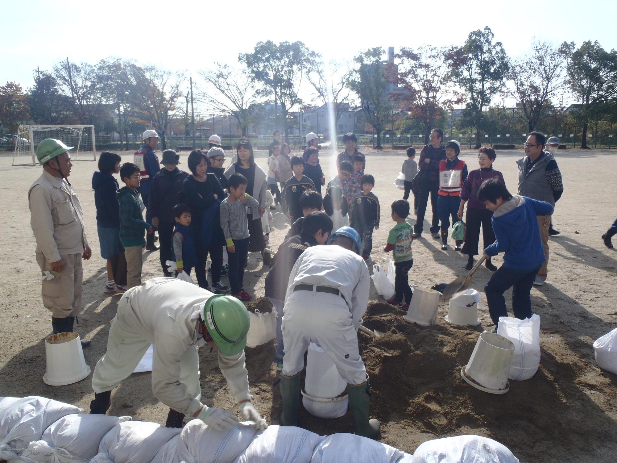 校庭で長久手緑化事業協力会の指導の下、準備された土をスコップですくって土のう作りをしている男の子たちを周りの参加者たちが見ている写真