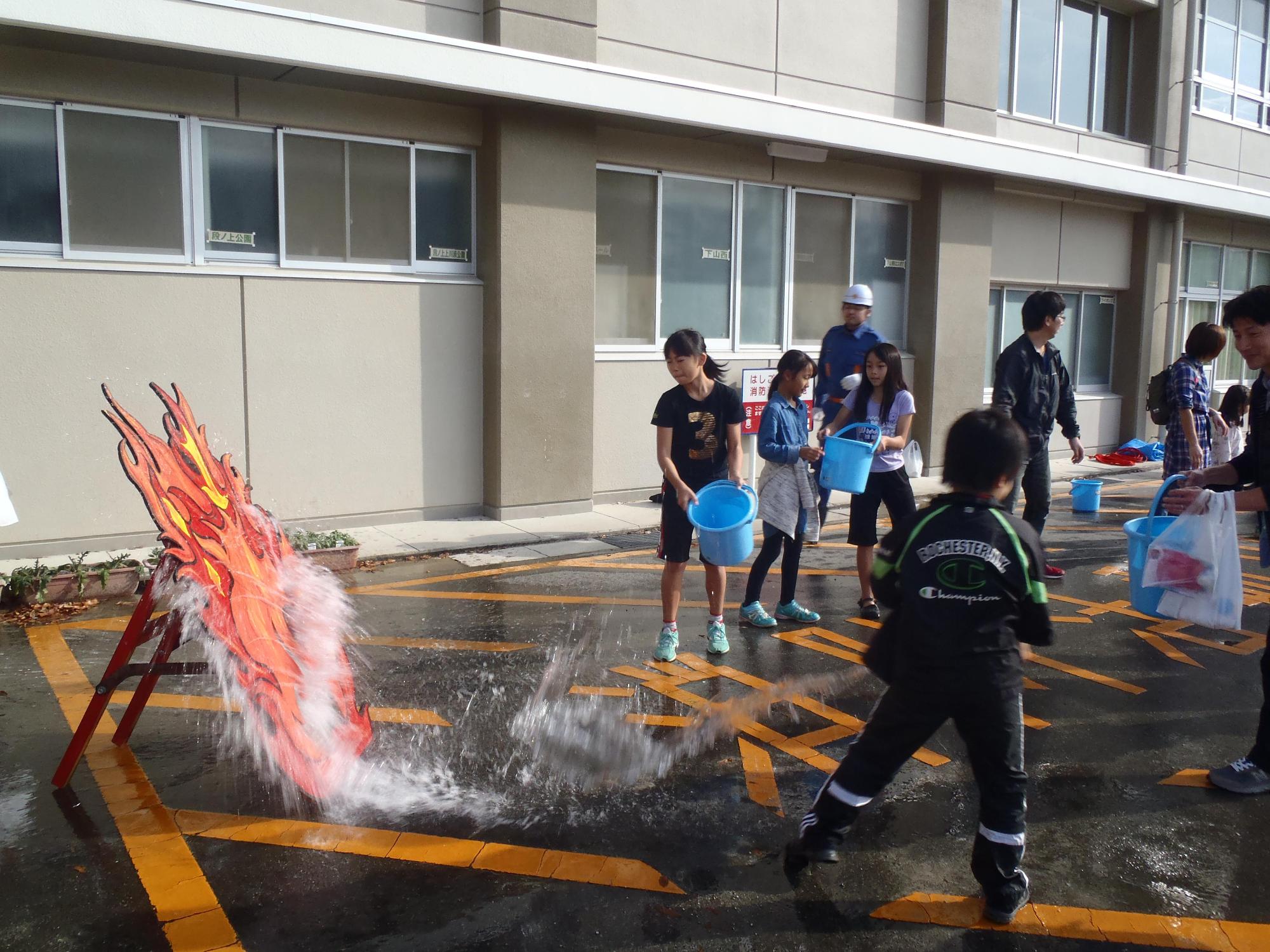消防団の指示の下、火のイラストに向けてバケツの水をかける子供たちの写真