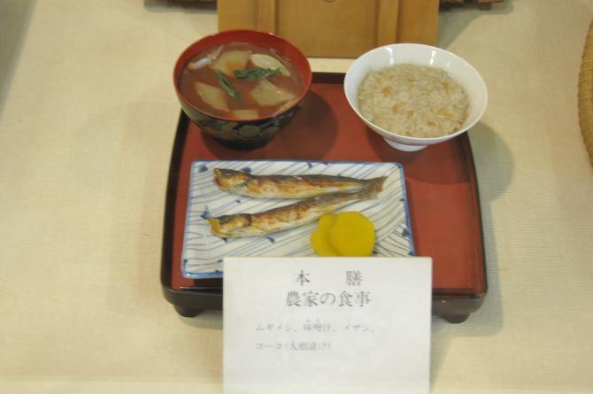 お膳台に味噌汁、麦飯、めざし2匹と沢庵が盛り付けられてある昭和初期の農家の食事の展示物の写真