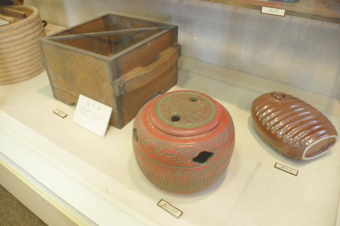 木製の四角い箱の形をした角斗舛、全体が朱色で緑色の模様が入っている陶器のねここたつ、茶色い湯たんぽが展示されてある写真