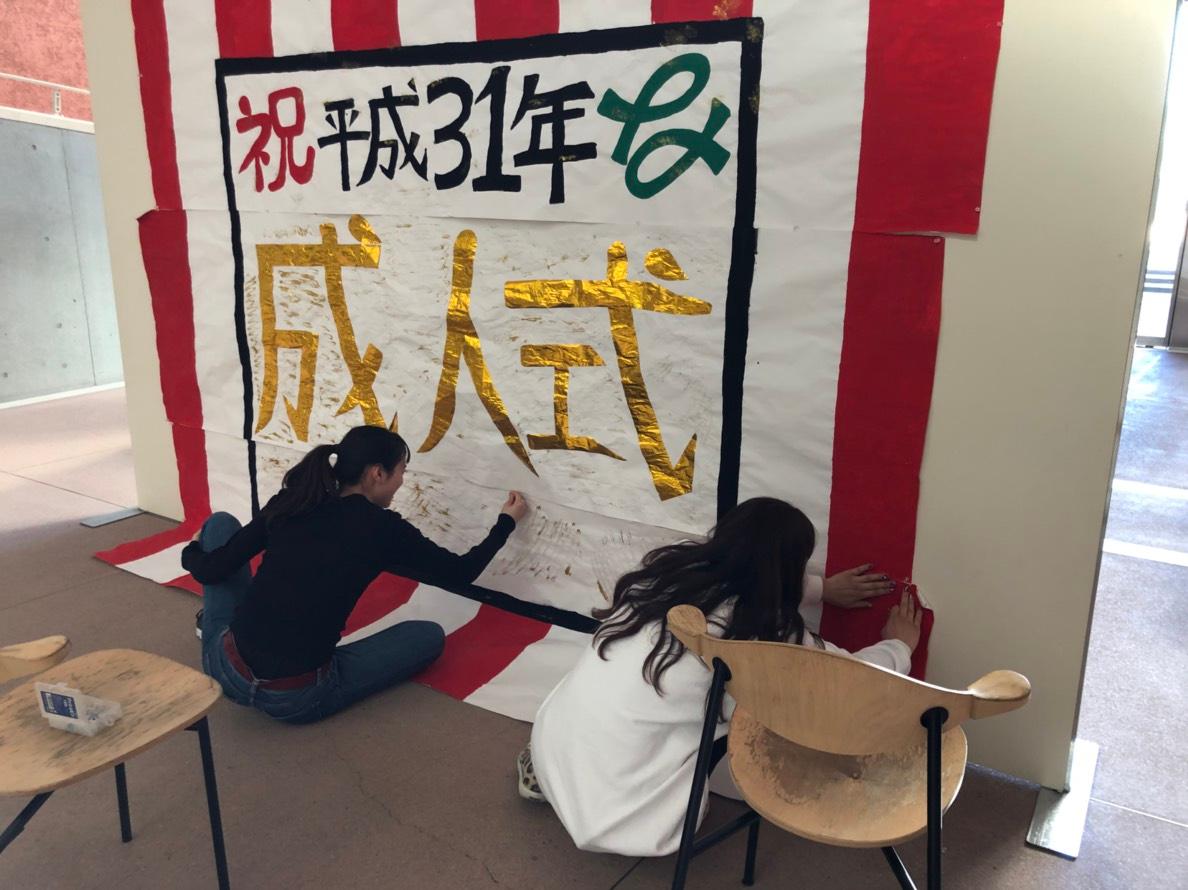 祝平成31年成人式と書かれた大きなパネルを作成している女性2人の写真