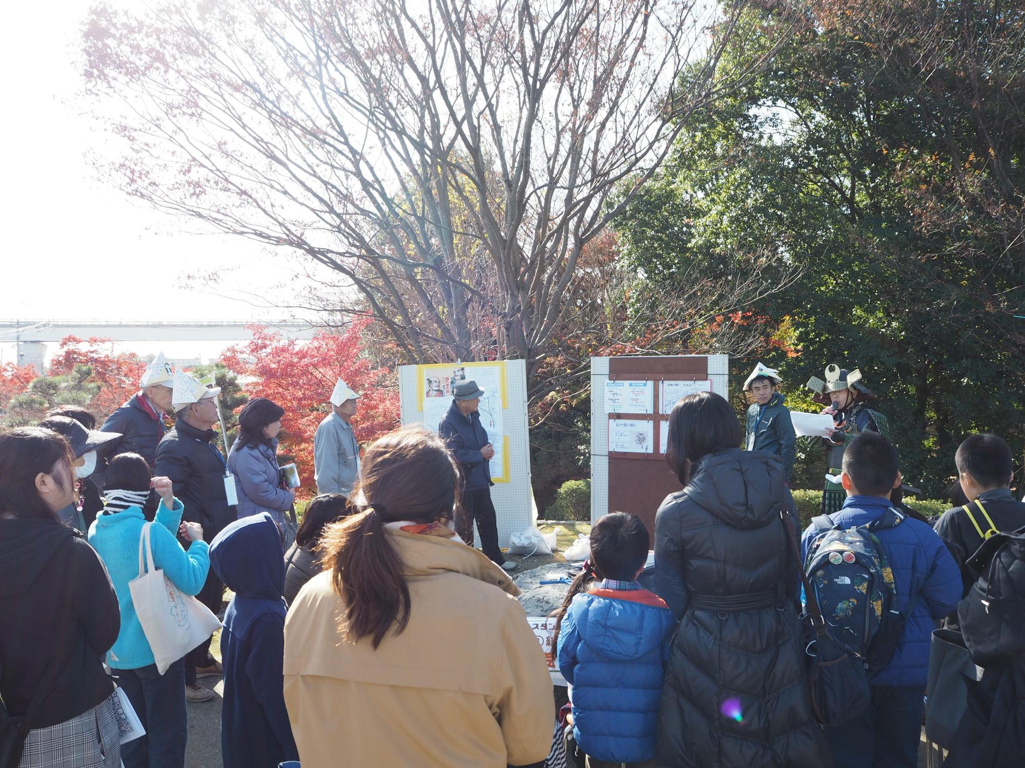 前方で紙芝居を読んでいる人の周りに大勢の人が集まっている写真