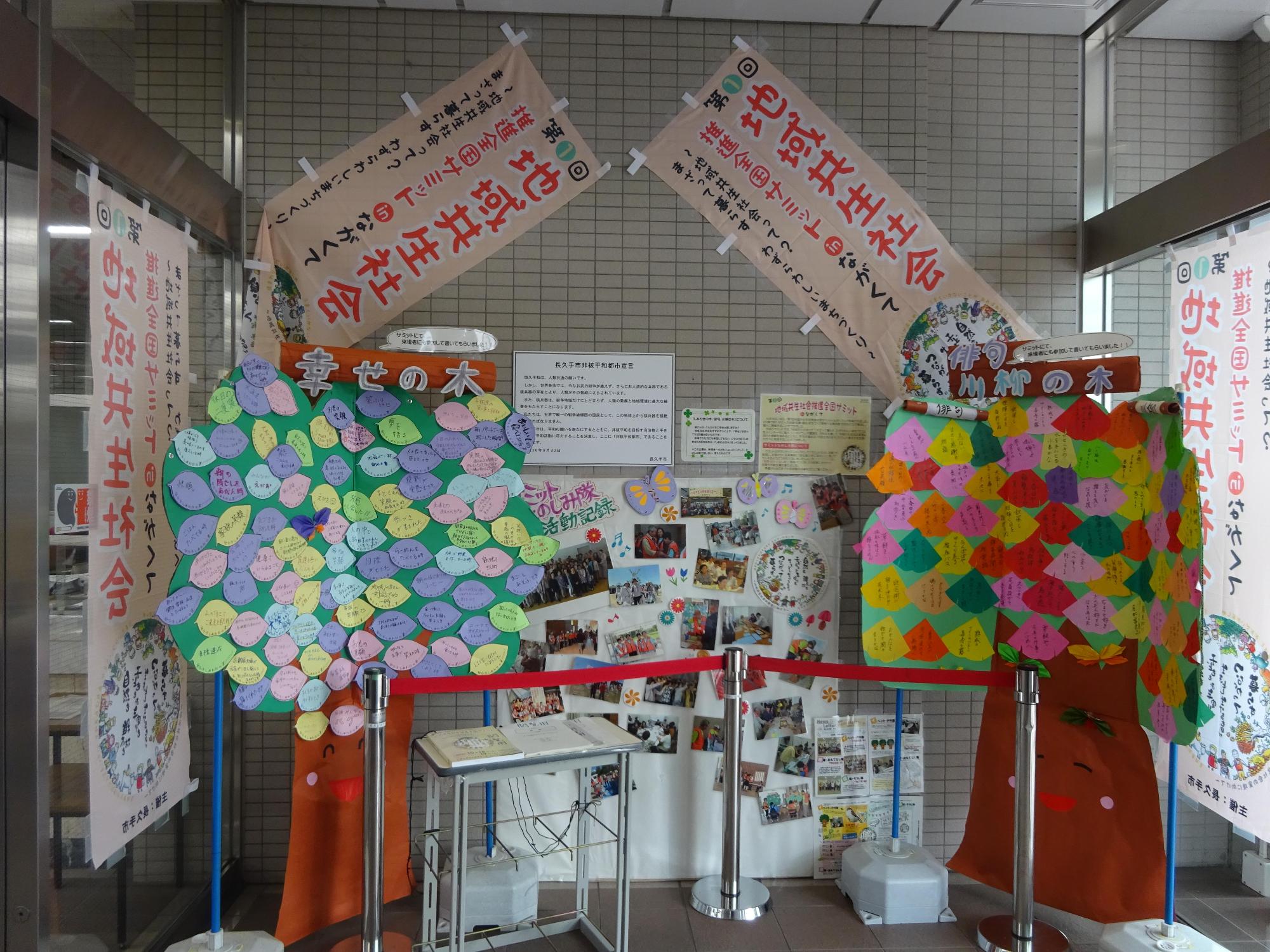 幸せの木と俳句川柳の木の間に模造紙に飾られた写真が展示され、地域共生社会ののぼり旗が展示されている玄関前の写真