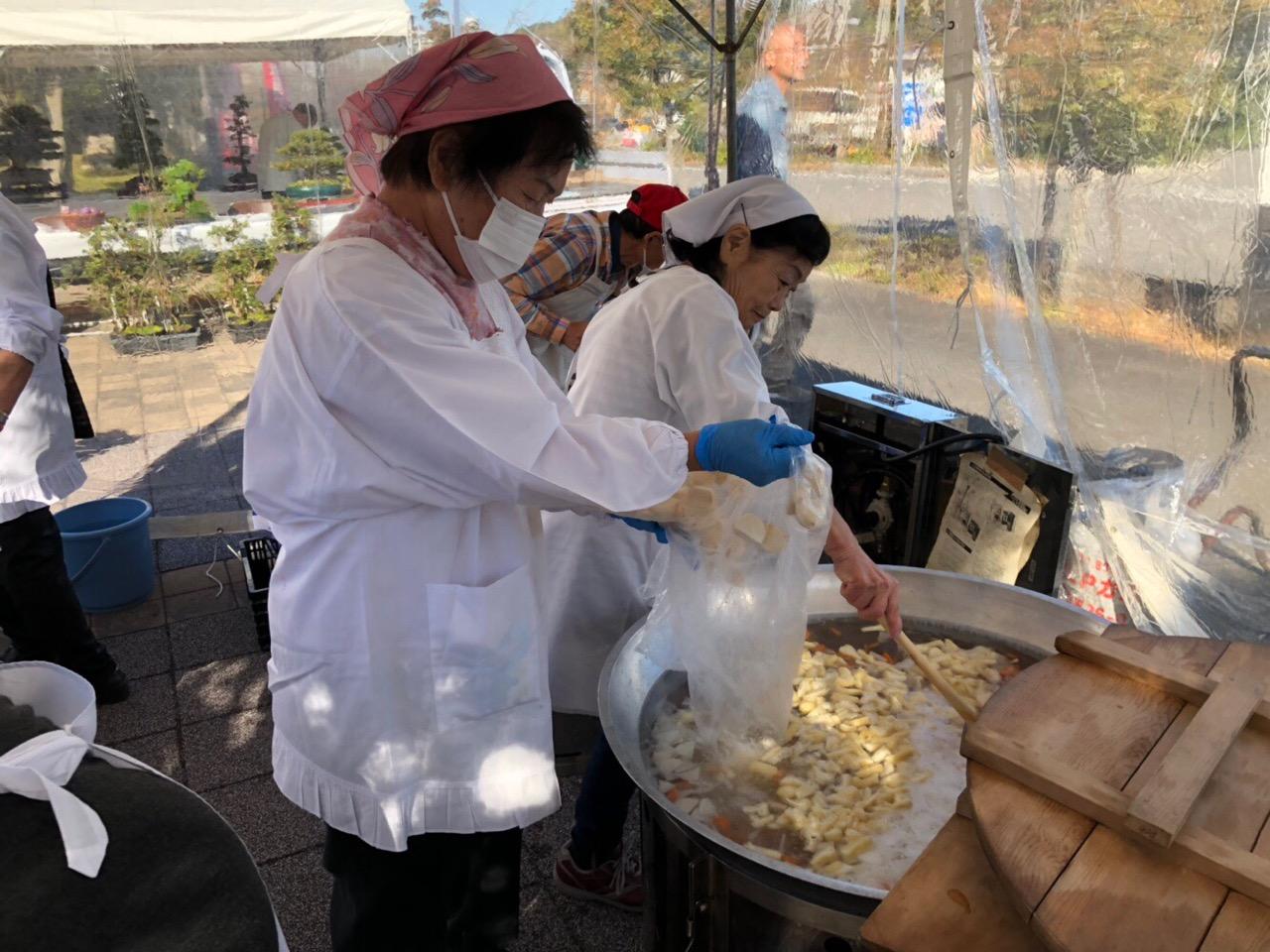 奉仕団の女性の方々が三角巾と割烹着を身に着けて、屋外のテントの中で、大鍋に汁物の材料を入れて煮込んでいる様子の写真