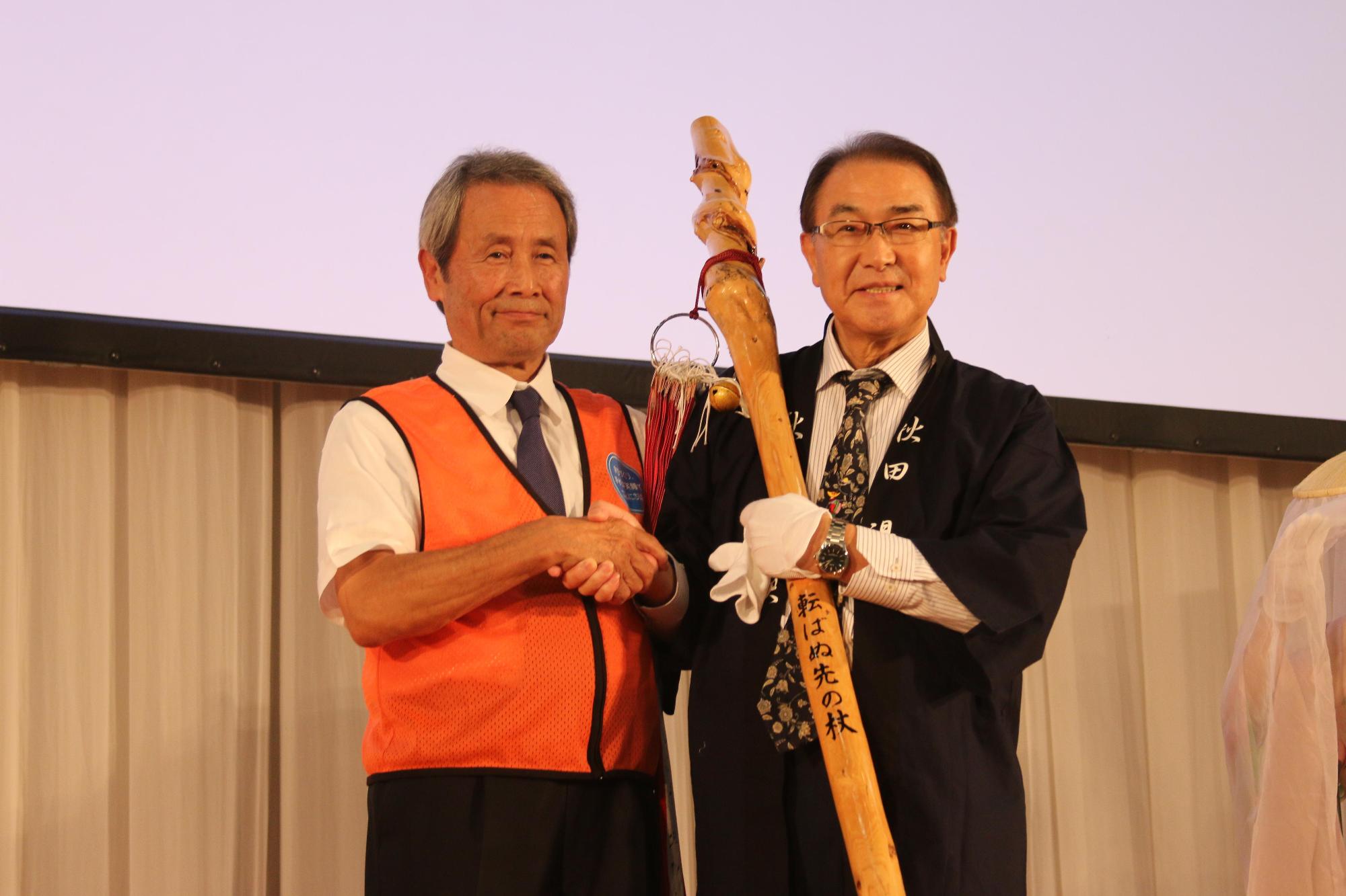 秋田県湯沢市の鈴木市長が「転ばぬ先の杖」を持って、吉田長久手市長と右手で握手をしている引継式の写真