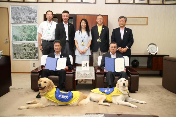 市長と大島慶久理事長が調印式の証書を胸の前に広げて椅子に座り、その前には介助犬2匹が座っており、関係者の方5名が後ろに並んで写っている写真