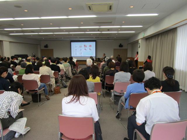 スクリーンに映し出された資料の前で講師が講演しているのを参加者が椅子に座って聞いている様子を後ろから撮影した写真