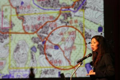 舞台上のスクリーンに映し出された地形図と講演をしている山村亜希さんの写真