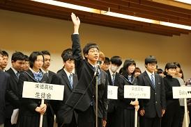 プラカードを持った女子生徒の後ろに各チームが並んでおり、代表の眼鏡をかけた男子生徒が、スタンドマイクの前で右手を挙げて選手宣誓をしている写真