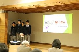 舞台上のスクリーンに資料が映されており、左端に4名の男子生徒が立っており、その中の右脚の男子生徒がマイクを持って話をしている写真