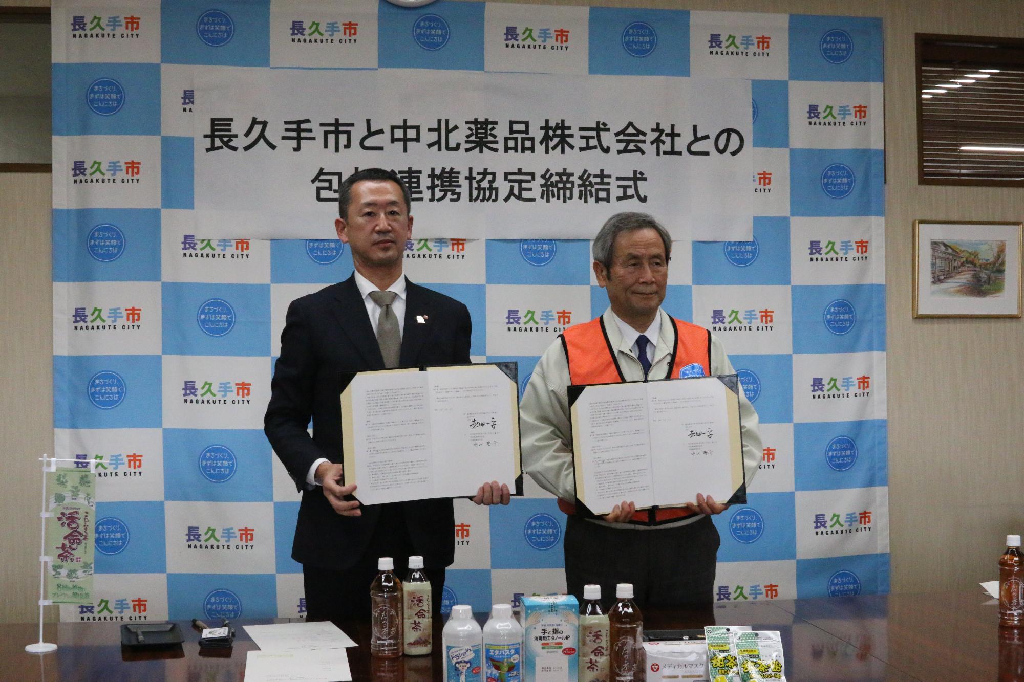 締結式にて中北薬品の商品が並べられているテーブルの後ろに立って長久手市長と中北薬品株式会社の代表がそれぞれ協定書を持って記念撮影している写真