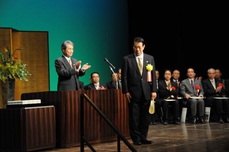 舞台上で表彰状をもって立っている加藤梅雄前町長に向かって拍手をしている吉田一平市長の写真