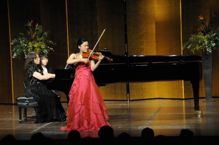 グランドピアノを演奏している2人の女性とバイオリンの演奏をしている赤いドレスの徳田真侑さんの写真