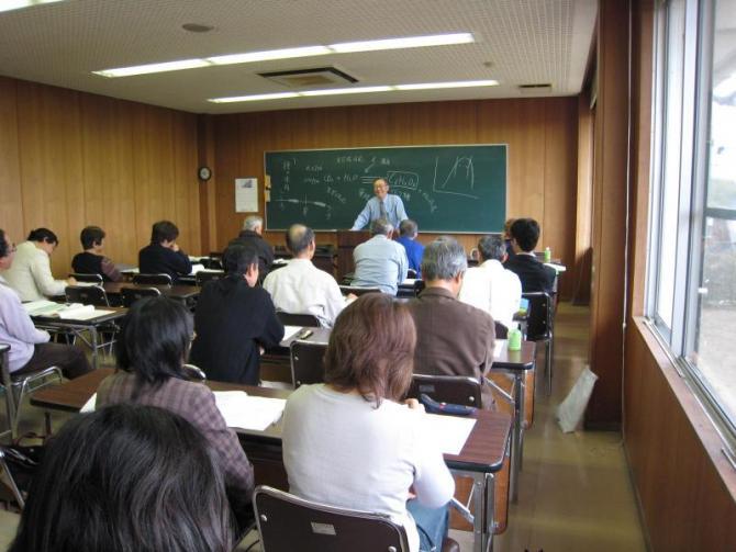 長机に生徒2名ずつ座っており、教室の黒板前に1名の男性が立ち、講義をしている写真