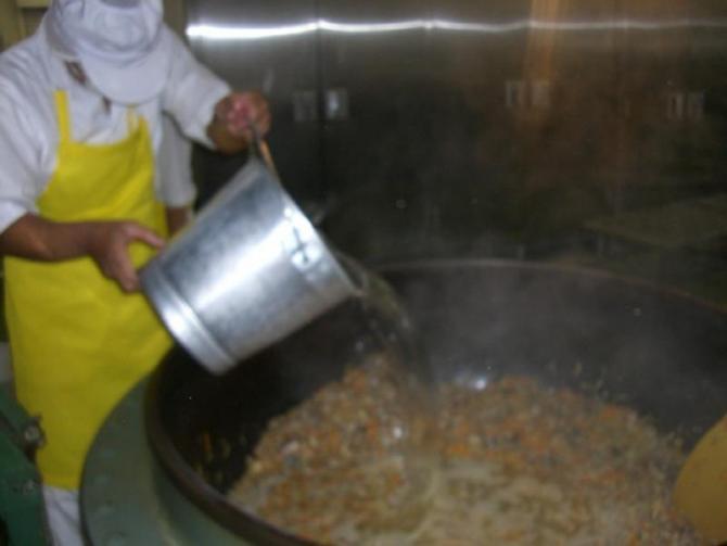 白い作業着と帽子を被り黄色のエプロンを着用したスタッフが、材料を炒めた大きな釜の中に銀色の食缶で水を大量に入れている写真