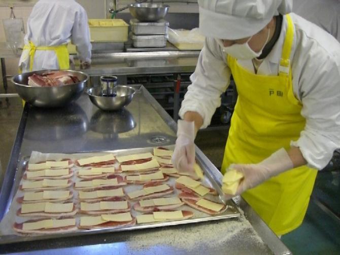 白い作業着に黄色のエプロンを着用したスタッフが、天板に豚肉を並べチーズを乗せている写真