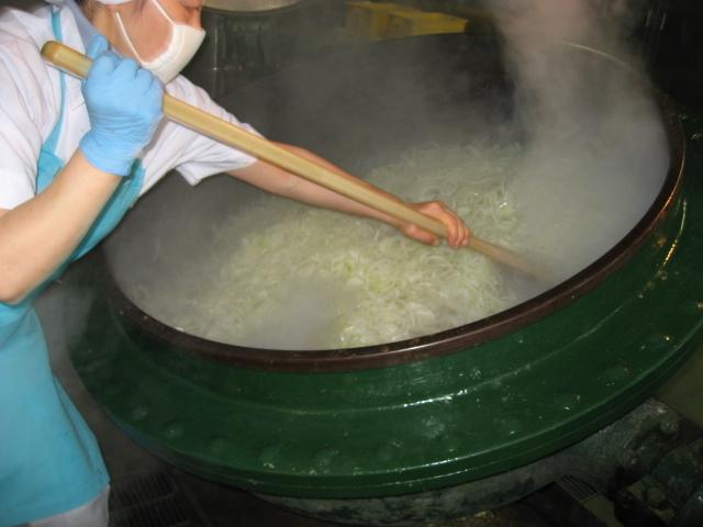 白い作業着に水色のエプロンを着用したスタッフが、大きな釜に入れた材料を炒めている写真