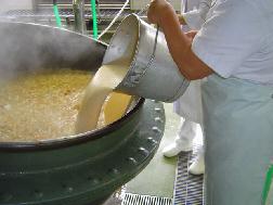 白色の作業着を着たスタッフが、大きな釜の中に食缶に入っている白い液体を大量に投入している写真