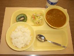 黄色のトレーに白米、スプーン、カレー、サラダ、キウイがのった給食の写真