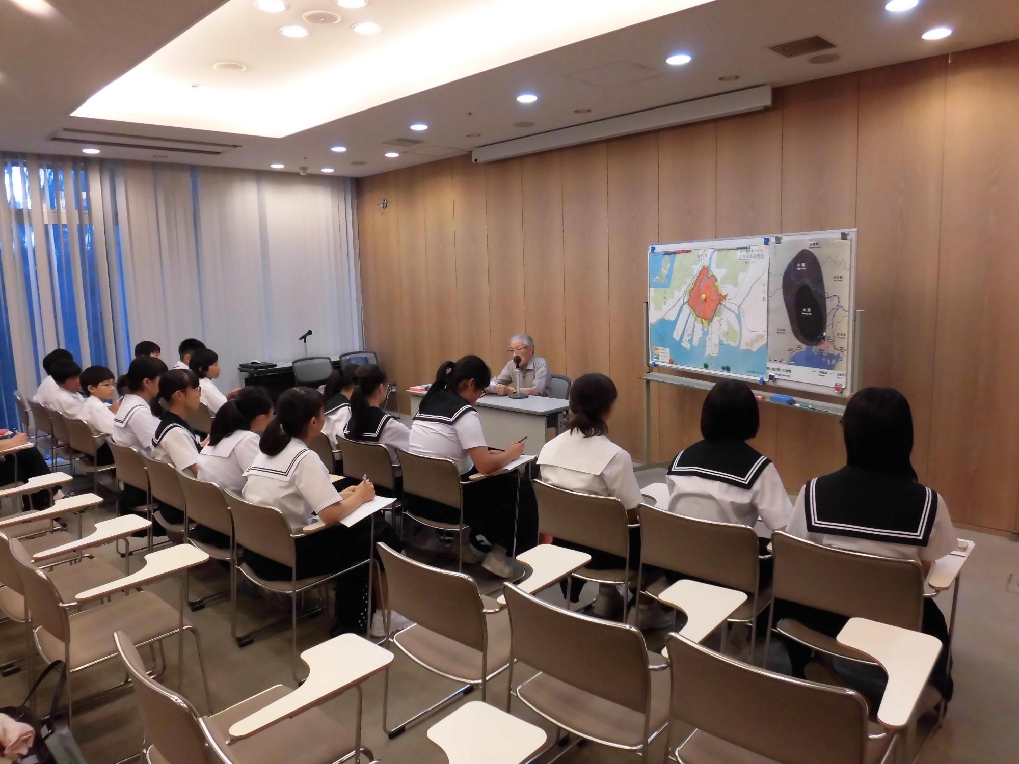浅野温生（あさのよしお）さんが前の机に座って体験談を話しており、生徒達が椅子に座ってメモを取りながら熱心に話を聞いている様子の写真