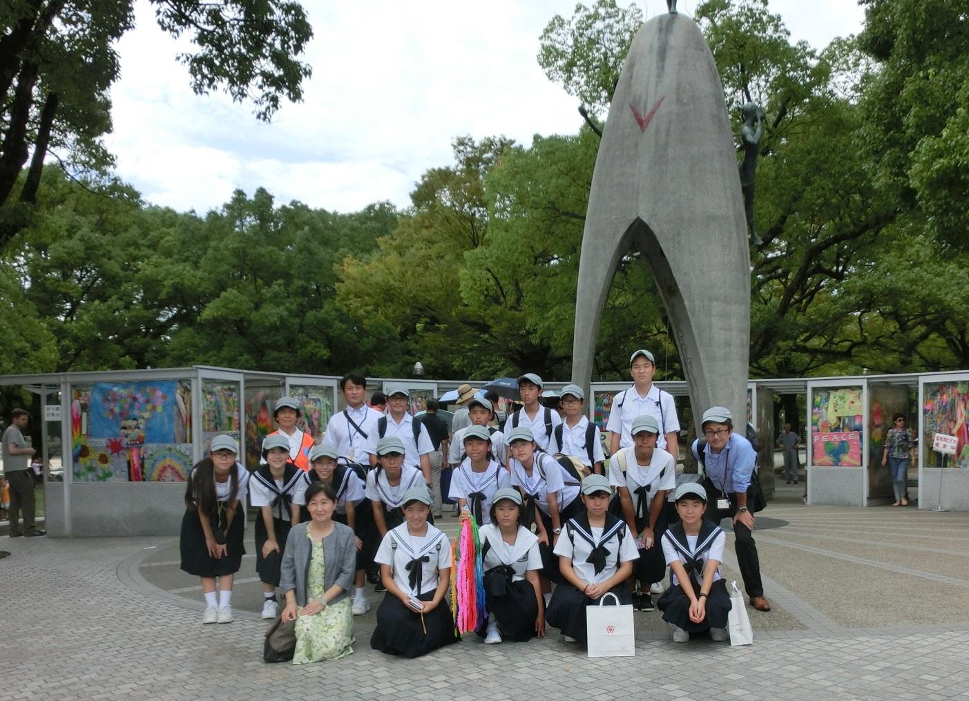 原爆の子の像の下で生徒達が笑顔で写っており、中央の女性生徒が千羽鶴を手に持って写っている集合写真