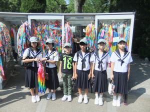 沢山の捧げられている千羽鶴の前で左端の女子生徒が千羽鶴を手に持って、女子生徒5名と男子生徒1名が並んで写っている写真